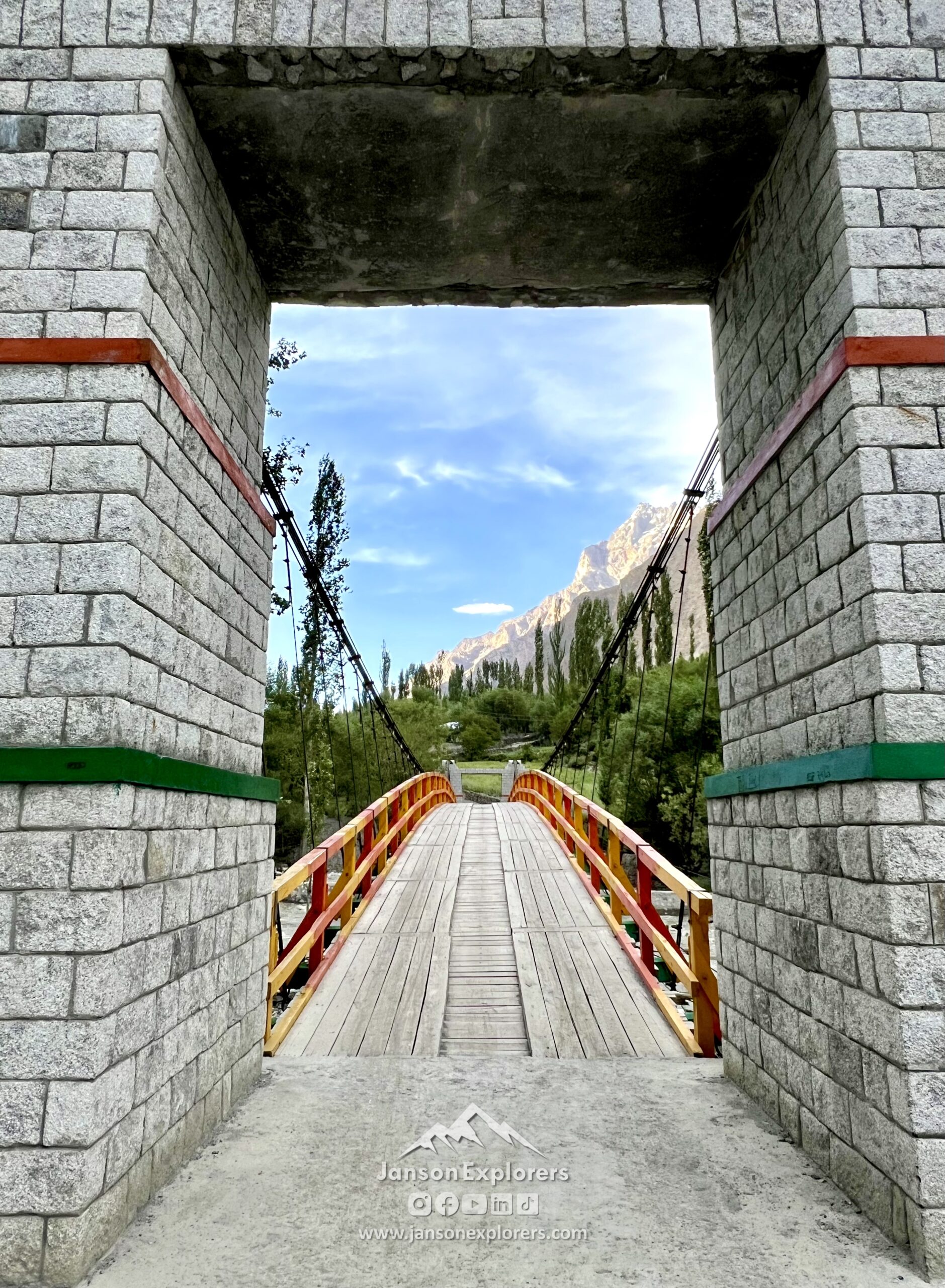Wooden suspension bridge framed by stone pillars with mountains and trees in Yaseen, Ghizer, Gilgit-Baltistan, Northern Pakistan