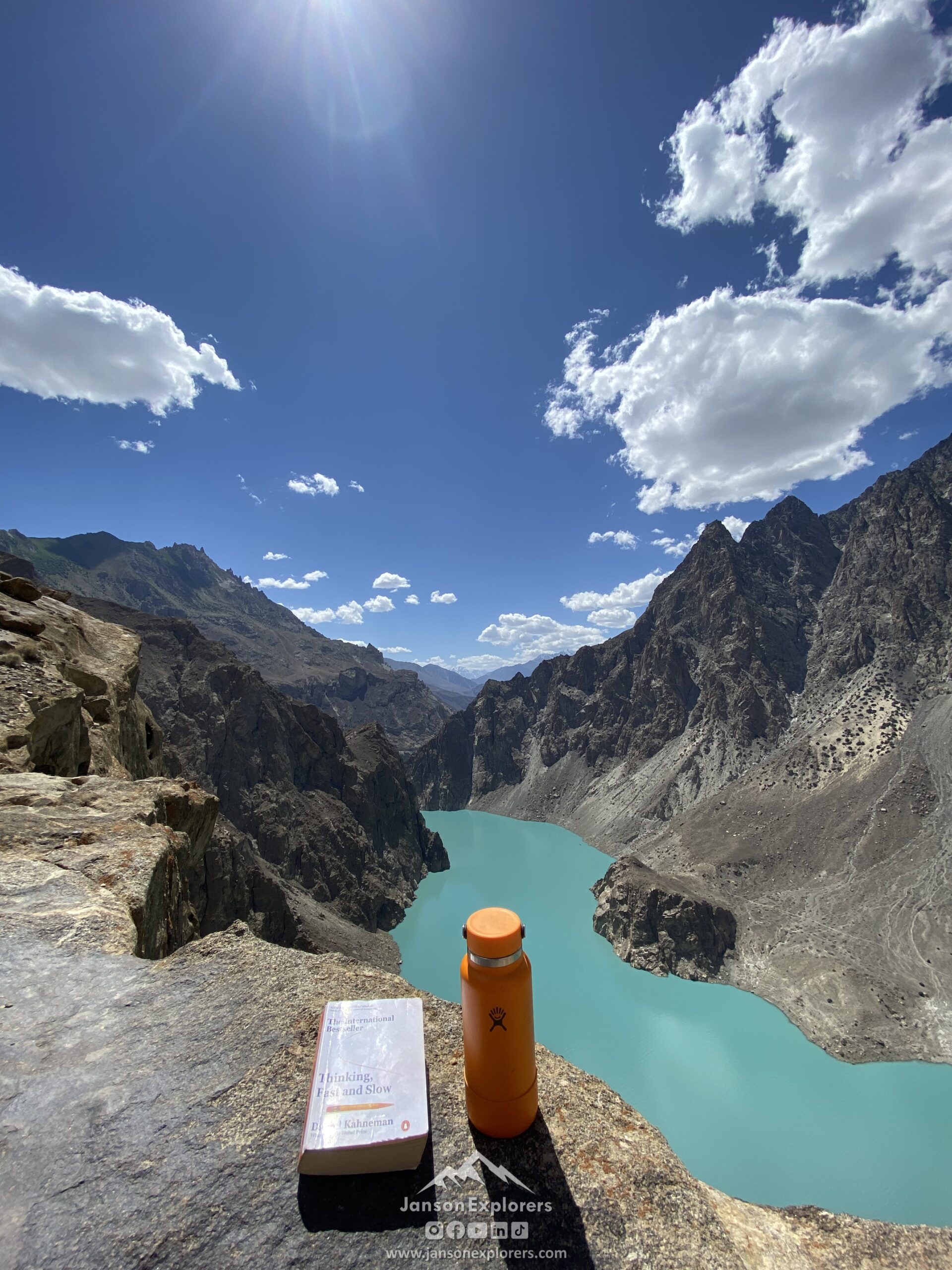 Hiking viewpoint above Attabad Lake, Hunza, with turquoise water winding between steep rocky mountains under a bright blue sky.